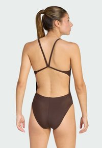 Una mujer joven con una coleta que lleva un traje de baño entero marrón, con la espalda baja y finas tiras cruzadas.