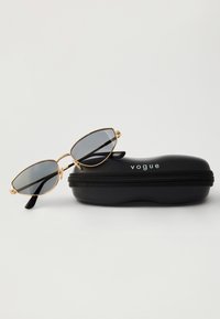 Sunglasevi u obliku mačjih očiju s zlatnim okvirom i tamnim staklima, odloženi na crnoj tvrdoj futroli sa zatvaračem, na kojoj je ispisana riječ "vogue."
