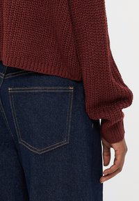 Burgunderfarget strikket genser med brede ermer parret med mørkeblå denimjeans, med gule sømmer og enkle baklommer.