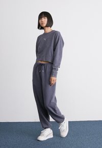 Sudadera gris recortada con un logo sutil, combinada con pantalones de chándal grises holgados. Zapatillas blancas completan el atuendo casual de athleisure.