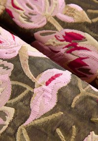 Bestickter Stoff mit großen rosa Blumenmustern auf einem dunkelgrünen Hintergrund, mit aufwendigen Details und einer strukturierten Oberfläche.