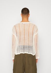 Man sett bakifrån i en vit, löst stickad långärmad tröja och olivgröna cargobyxor mot en enfärgad bakgrund.