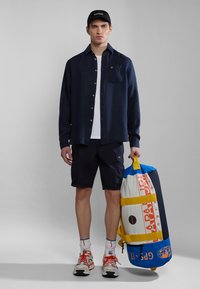 Camicia blu con bottoni, t-shirt bianca, pantaloni cargo blu, sneakers grigie con dettagli arancioni, mentre tiene una borsa a tracolla multicolore.