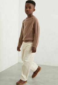 Brun stickad tröja med långa ärmar, krämfärgade byxor och bruna slip-on skor. Står på en grå golv mot en ljus bakgrund.