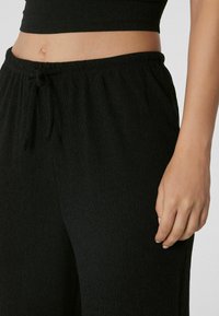 Pantalons noirs texturés avec une taille élastique, dotés d'un cordon de serrage et d'une coupe ample, présentés au niveau de la taille et des mains.