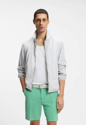 Jeune homme portant une veste blanche à fermeture éclair, un débardeur blanc, un short vert et une ceinture de couleur claire, debout les mains dans les poches.