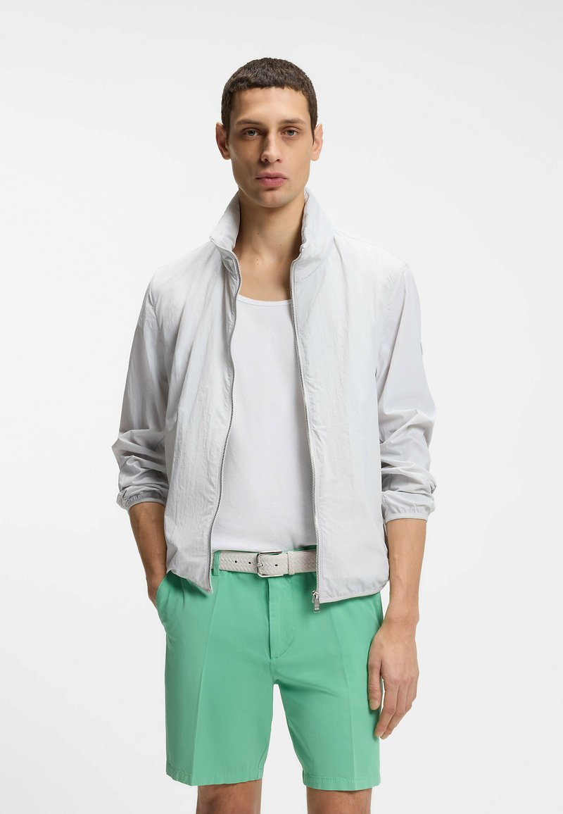 Jeune homme portant une veste blanche à fermeture éclair, un débardeur blanc, un short vert et une ceinture de couleur claire, debout les mains dans les poches.