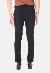 Uomo che indossa pantaloni di velluto a coste neri e scarpe eleganti nere, in piedi con le braccia rilassate ai lati, di spalle rispetto all'osservatore su uno sfondo bianco.