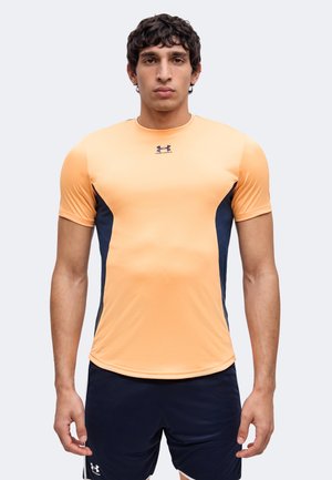 Jonge man met een perzikkleurig en marineblauw sportshirt met korte mouwen en marineblauwe korte broek, staand met een neutrale gezichtsuitdrukking recht vooruit kijkend.