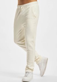 Kremfargede sweatpants laget av myk stoff har snøring i livet, sidelommer og subtil brodering på hoften, sammen med hvite joggesko.