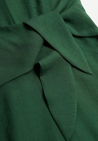 Tissu vert avec une texture lisse mettant en avant un détail de nœud prononcé et des plis. Les bords présentent une couture contrastante pour une définition accrue.