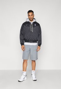 Musta satiin bomber-takki, jossa on NBA-logo, harmaa huppari alla, harmaat sortsit ja valkoiset lenkkitossut mustilla yksityiskohdilla.