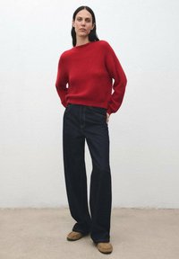 Donna con lunghi capelli scuri in piedi indossando un maglione rosso, jeans larghi scuri e scarpe mocassino beige contro un muro bianco semplice.