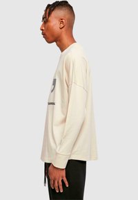 Langarm-Hemd aus leichtem beigen Baumwollstoff mit Rundhalsausschnitt, auf der Vorderseite mit einem grauen Grafik- und Textdruck. Lockere Passform mit fallenden Schultern.