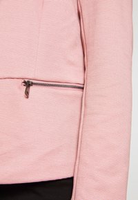 Ljusrosa jacka med en strukturerad, randig mönstring, försedd med en svart dragkedja och en stilren, figurnära design. Slät tyg och skräddarsydd silhuett.