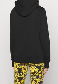 Felpa nera con cappuccio e vestibilità rilassata. Caratterizzata da polsini e orlo a coste. Abbinata a leggings gialli e neri con motivo barocco.