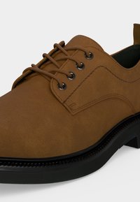 Chaussure en daim marron à lacets, semelle noire, œillets métalliques et surpiqûres visibles sur le côté et autour des lacets.