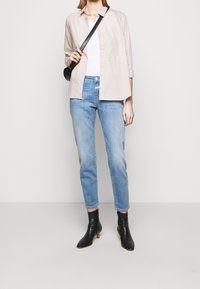 Camisa de botones color beige claro sobre una blusa blanca, combinada con jeans ajustados azul claro y botas negras hasta el tobillo. Acento de bolso cruzado negro.