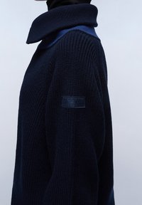Granatowy sweter dzianinowy z wysokim, złożonym kołnierzem i prążkowaną teksturą. Posiada małą naszywkę z logo marki na rękawie. Widok z boku.