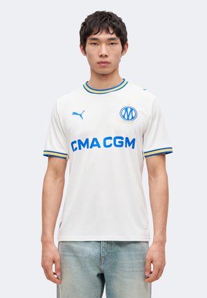 Giovane uomo che indossa una maglia da calcio bianca del Marsiglia con rifiniture blu e gialle, logo CMA CGM e emblema Puma, in piedi davanti a uno sfondo semplice.
