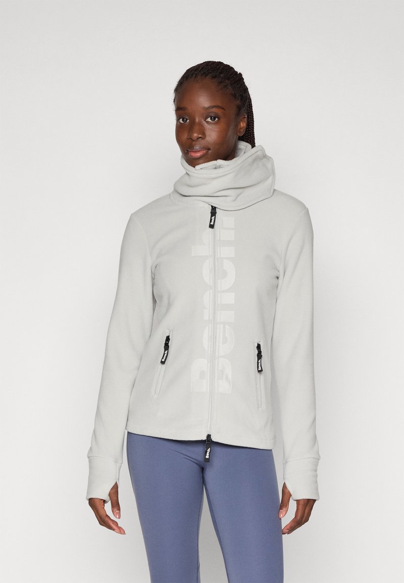 Lichtgrijze fleece jas met een hoge kraag, ritszakken en een groot merkenlogo aan de voorkant; zachte textuur en lange mouwen.