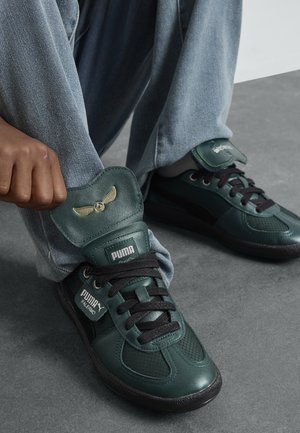 Zapatillas de cuero verde con detalles en negro, que incluyen una lengüeta acolchada, lados texturizados y un emblema dorado en la lengüeta. Cintas negras.