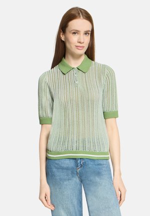 Młoda kobieta w zielono-białej, paskowanej dzianinowej koszulce polo z krótkim rękawem i niebieskich dżinsach, stojąca na białym tle.