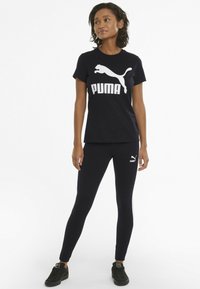 Czarna bawełniana koszulka z białym logo Puma w połączeniu z czarnymi legginsami z małym białym logo Puma. Model ma na sobie czarne sneakersy.