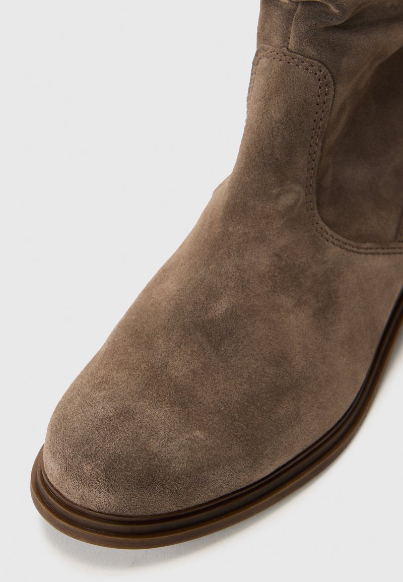 Botín de ante marrón con punta redonda y detalles de costura, que presenta una textura suave y una suela marrón contrastante.