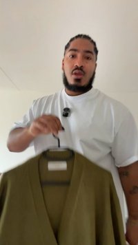 Oliivinvihreä neulottu neuletakki, V-kaula-aukolla, kevyellä tekstuurilla ja resorikuvioinneilla, mustalla henkarilla ripustettuna, henkilön pitämänä.