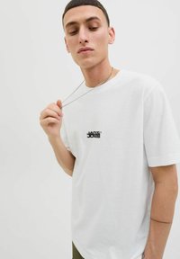 White cotton t-shirt with short sleeves, featuring a black logo centered on the chest. The model wears a silver chain necklace.