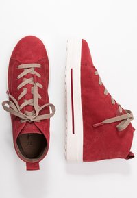 Czerwone zamszowe wysokie trampki z beżowymi sznurowadłami, białą gumową podeszwą i czerwonym paskiem jako akcentem. Posiadają zaokrąglony czubek i tylną pętelkę.