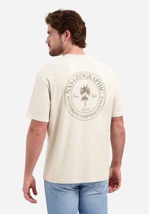 Man met bruin haar die een beige T-shirt draagt met een palmboomafbeelding en de tekst "NXS Geographic Mysterious Impermanence" op de rug.