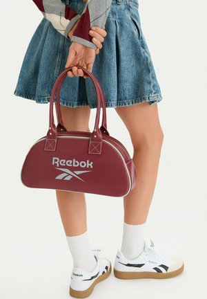 Person in Denimrock und Rautenpullover hält eine kastanienbraune Reebok-Handtasche, trägt weiße Reebok-Sneaker und weiße Socken vor einfarbigem Hintergrund.