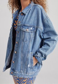 Blå denimjacka med en avslappnad passform, som har två bröstfickor, kopparknappar och slitage detaljer längs fållen och ärmarna.