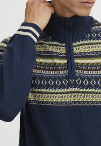 Kékeszöld pulóver magas gallérral, geometrikus mintákkal zöld, barna és fehér színekben. Cipzáros záródás a nyaknál. Csíkos mandzsetta.