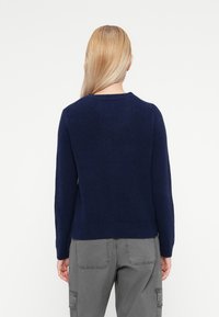 Pulover tricotat de culoare bleumarin, cu gât rotund și tiv texturat, văzut din spate. Material moale, texturat, combinat cu pantaloni cargo gri.
