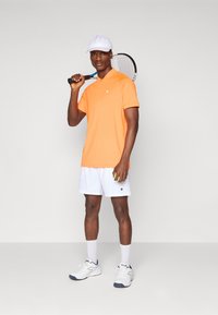 La tenue de tennis comprend un polo orange à manches courtes et un short blanc, associé à une casquette blanche et des baskets. Tient une raquette de tennis et une balle.