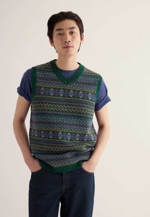 Giovane uomo con le mani in tasca indossa maniche blu arrotolate e un gilet in maglia verde a fantasia, su uno sfondo chiaro e uniforme.