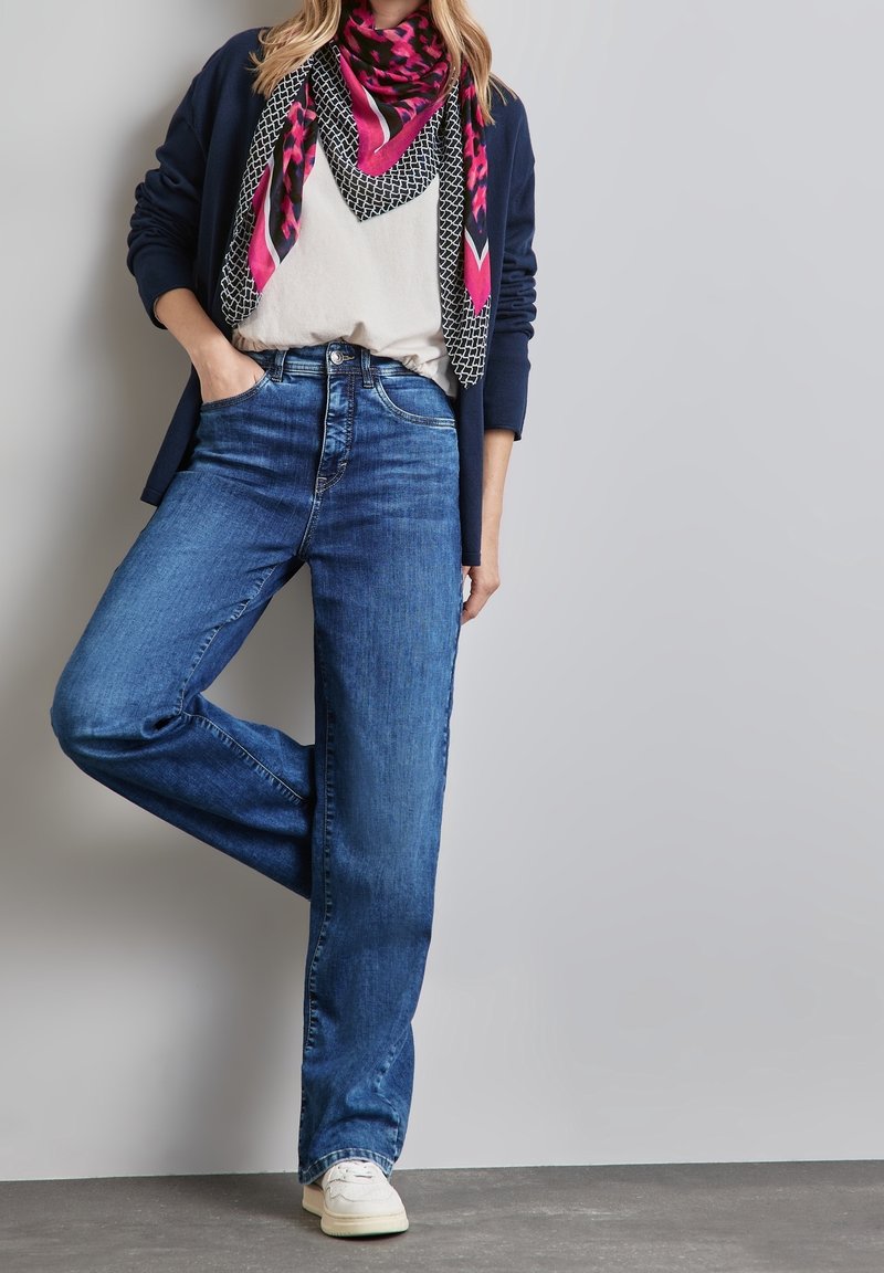 Femme en tenue décontractée avec jean bleu, baskets blanches, haut blanc, cardigan bleu marine et écharpe rose à motifs sur fond gris uni.