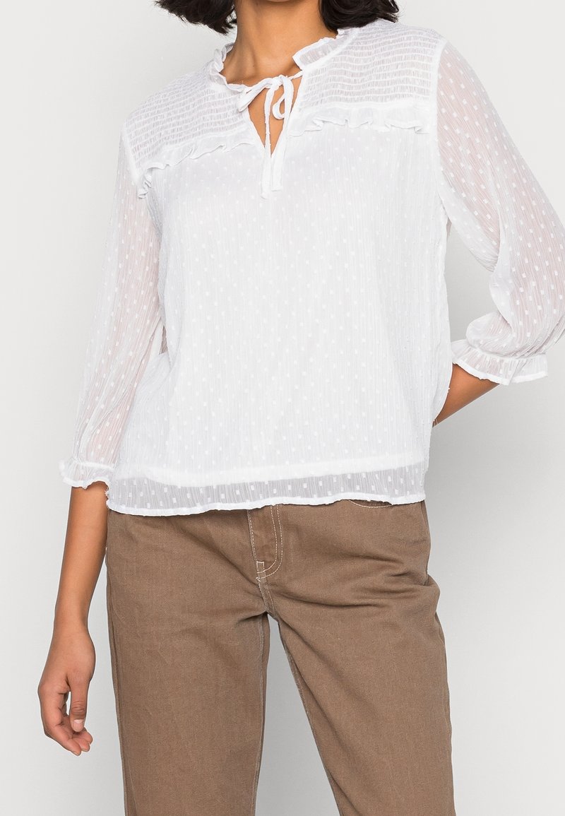White blouse with textured polka dot pattern, ruffled neckline, and tie detail. Sheer long sleeves with elastic cuffs, paired with brown pants.