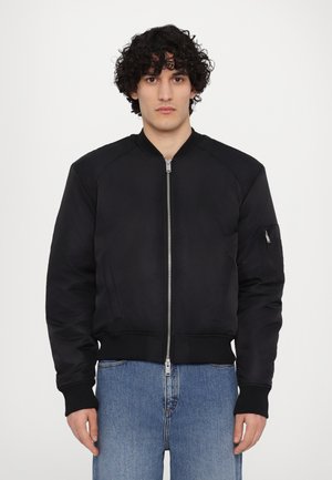 Melna bomber jaka ar rāvējslēdzēju priekšā, ribotām piedurknēm un apakšmalu, izceļot labās rokas kabatu ar rāvējslēdzēju. Gluda, viegla auduma struktūra.