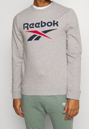 Mężczyzna w jasnoszarej bluzie Reebok z granatowo-czerwonym logo, do tego jasnookie zielone spodnie z częściowym białym logo.