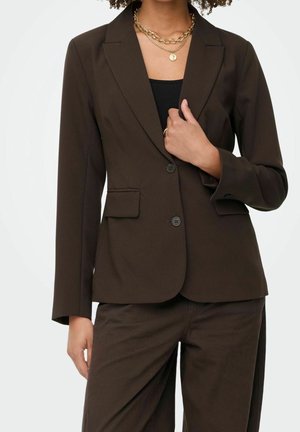 Vrouw draagt een donkerbruine, op maat gemaakte blazer met twee knopen, bijpassende broek, zwarte top en gelaagde gouden kettingen.