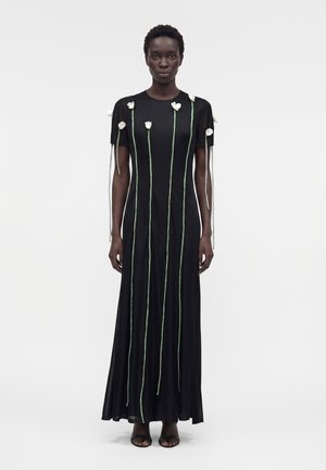 Frau trägt ein langes schwarzes Kleid mit vertikalen grünen Linien und weißen Blumenverzierungen auf Brust und Ärmeln, steht vor einem einfarbigen Hintergrund.