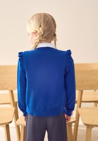 Maglione blu lavorato a maglia con dettagli arricciati sulle spalle, scollo rotondo e polsini aderenti, abbinato a pantaloni grigio scuro. Sullo sfondo si vedono sedie di legno.