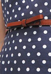 Marinblå klänning i tyg med vita prickar, med ett brunt läderbälte som har en rosett. Slät textur, figurnära design.