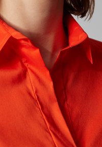 Czerwona koszula z kołnierzykiem o gładkiej, warkoczowej fakturze, z dopasowanym dekoltem i subtelnymi detalami przeszyci na kołnierzyku oraz listwie guzikowej.