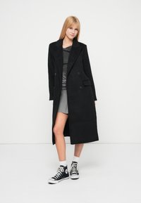 Palton lung negru, dublu cu revere mari, material texturat și buzunare laterale, asortat cu un top gri și adidași negri.