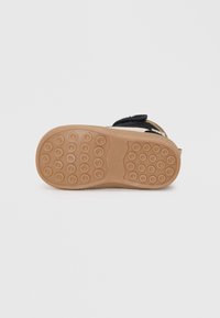Suola in gomma marrone con tasselli circolari, tomaia in tessuto nero con cinturino in Velcro, accenti beige lungo i bordi, design piatto, stile casual.